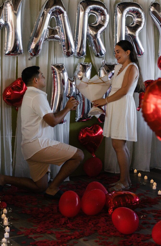 A heartfelt marriage proposal scene with balloons, candles, and rose petals creating a romantic atmosphere.